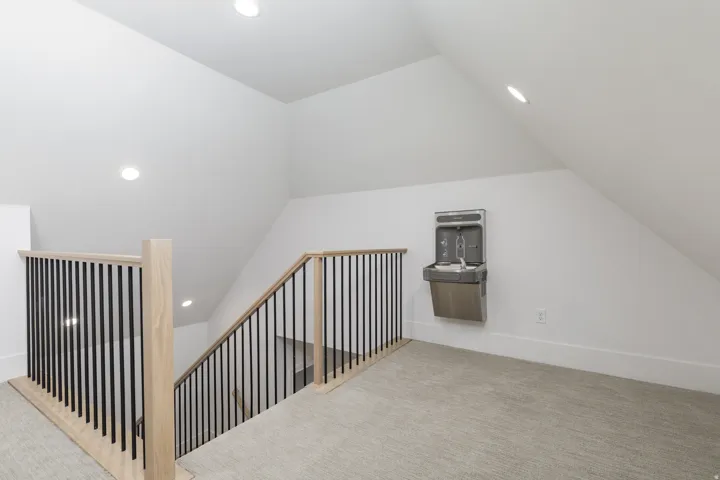 Carpeted loft area featuring a modern wood and metal staircase railing, recessed lighting, and a wall-mounted water dispenser