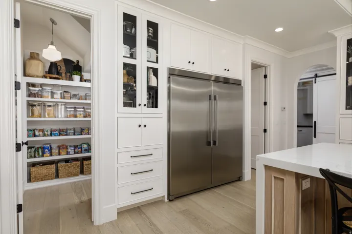 Expansive pantry with built-in shelving, white cabinetry with glass inserts, stainless steel refrigerator, light wood-finish flooring, and a kitchen island with a light-toned countertop