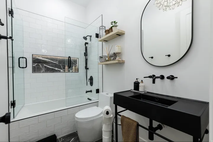 Contemporary bathroom featuring a tub-shower combination with glass enclosure, a built-in shower niche with stone-finish tile, a wall-mounted vanity with integrated sink, and an oval framed mirror