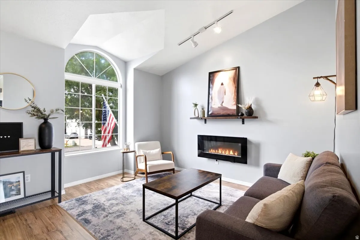 Vaulted ceilings, a large arched window, wood-finish flooring, a wall-mounted electric fireplace, and track lighting