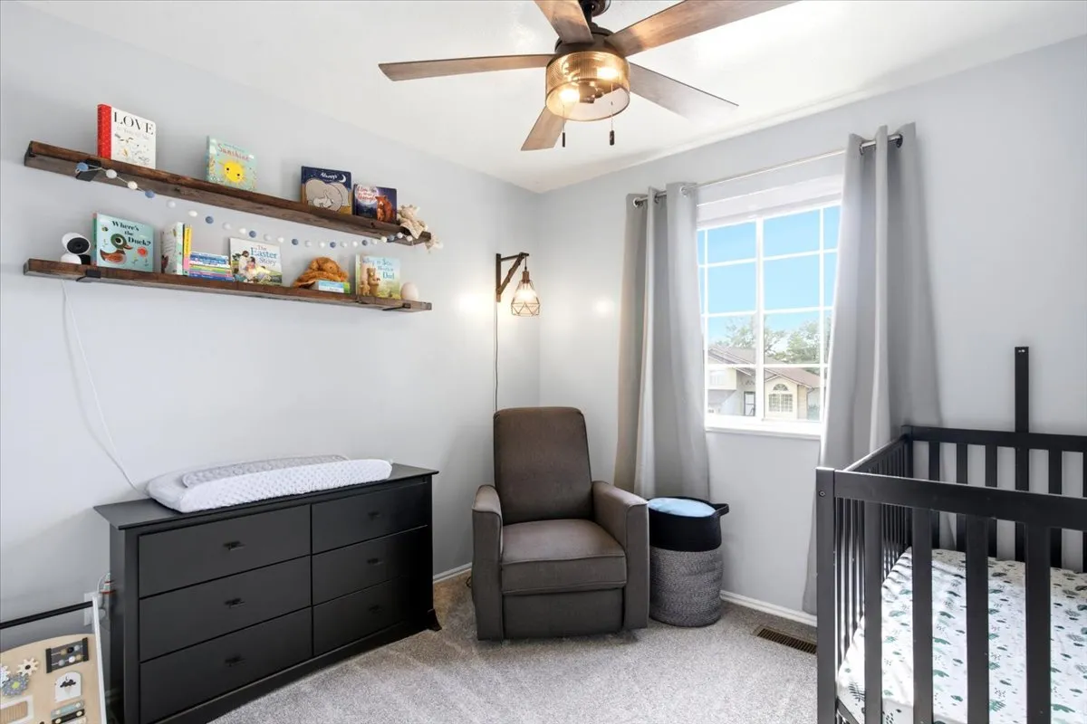 Neutral-toned wall paint, two wall-mounted wood-finish floating shelves, ceiling fan with integrated lighting, window with white trim, and light gray carpet flooring