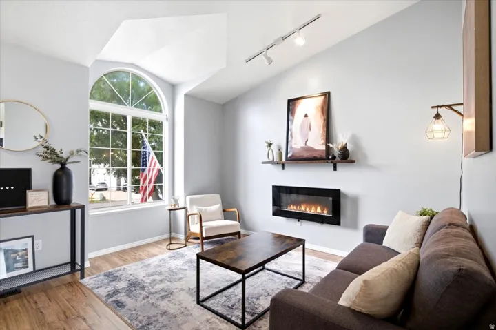 Vaulted ceilings, a large arched window, wood-finish flooring, a wall-mounted electric fireplace, and track lighting