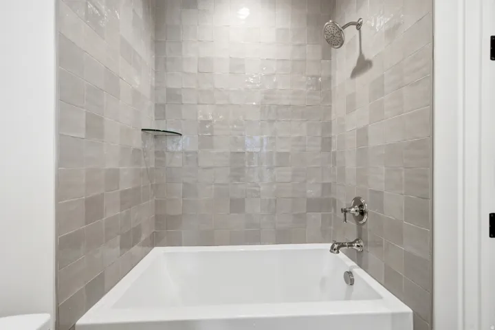 Bathtub and shower combination featuring tiled surround, chrome fixtures, and a clear glass shelf