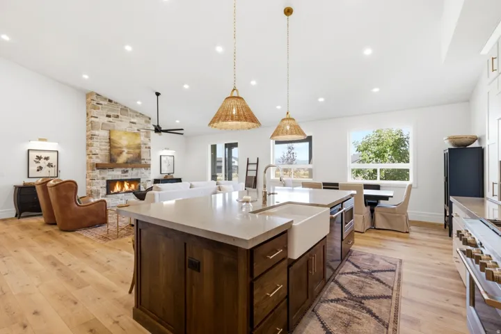 Expansive open-concept living area featuring a stone-clad fireplace, wood-finish flooring, recessed lighting, and an island with an integrated sink and light-toned countertop