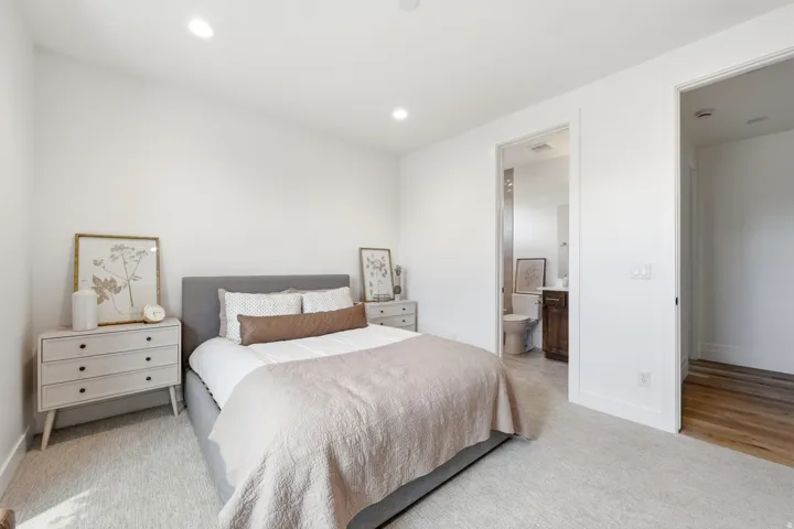 Recessed lighting, light-colored carpeting, white wall finishes, and a doorway leading to a bathroom with a dark wood-finish vanity
