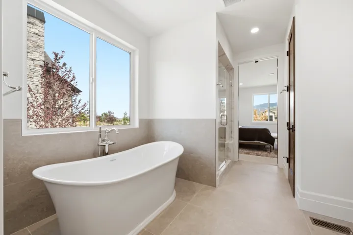 Freestanding soaking tub with chrome fixtures, large window, glass-enclosed shower, and neutral tile flooring