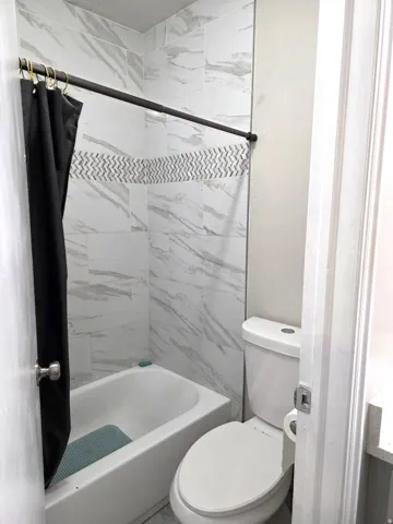Bathtub with a shower, white tile surround, decorative mosaic tile accent, and a white toilet