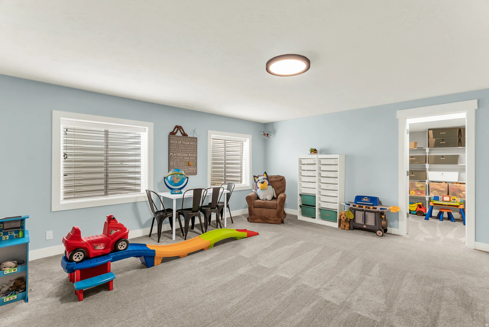 Recessed lighting, light blue wall paint, two windows with blinds, carpeted flooring, and a doorway leading to an additional space with built-in shelving