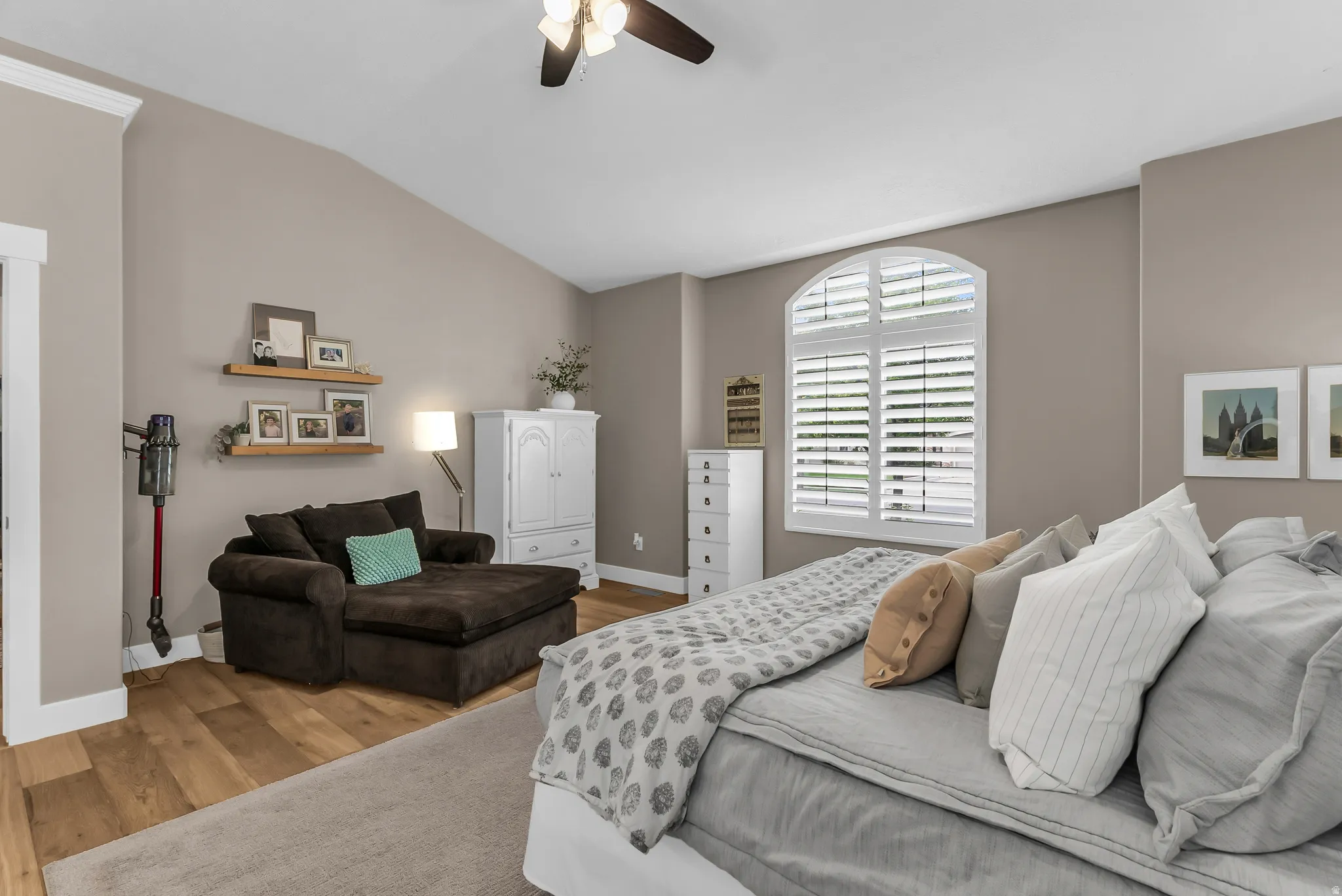 Vaulted ceiling, wood-finish flooring, arched window with plantation shutters, ceiling fan, and floating wall shelves