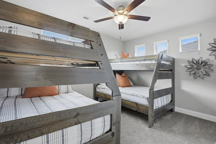 Two wood-finish bunk beds with integrated ladders, ceiling fan with light fixture, three high-set windows, and light gray wall paint
