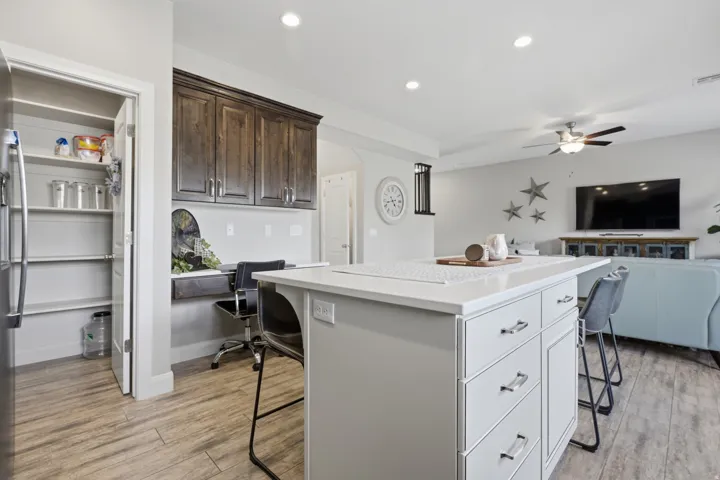 Spacious open-concept layout featuring a white kitchen island with an integrated breakfast bar, wood-finish flooring, recessed lighting, and a built-in desk area with dark wood cabinetry
