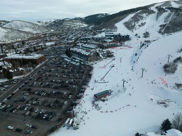 Expansive ski area with multiple chairlifts and slopes, surrounded by snow-covered mountains, and a parking area with numerous vehicles