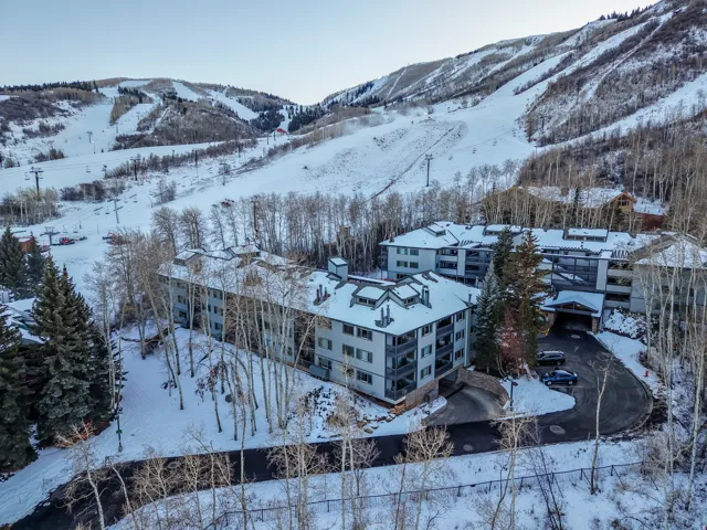 Multi-story residential buildings with balconies, surrounded by mature trees, situated on a snow-covered landscape with visible ski slopes
