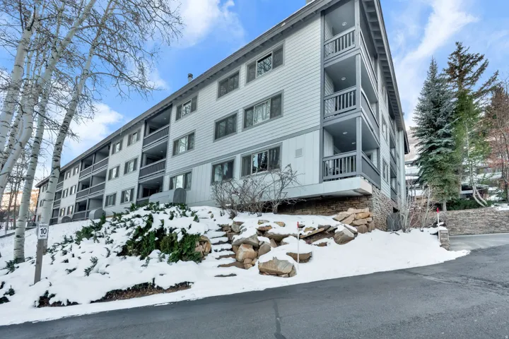 Multi-story building featuring balconies with railings, sloped terrain with rock accents, and mature trees surrounding the property