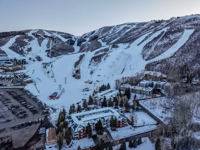 Snow-covered mountain terrain with ski slopes, multiple structures with varying architectural styles, snow-covered trees, and a visible parking area