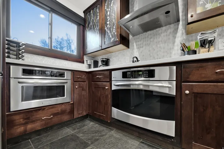 Stainless steel built-in appliances, dark wood cabinetry, light-toned countertops, and a patterned tile backsplash complement the dark tile flooring