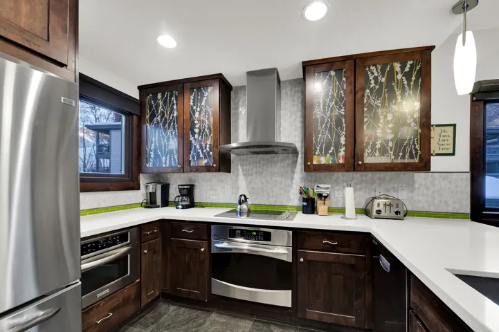 U-shaped kitchen with dark wood cabinetry, white countertops, built-in stainless steel appliances, a stainless steel range hood, and a textured backsplash with a green accent tile