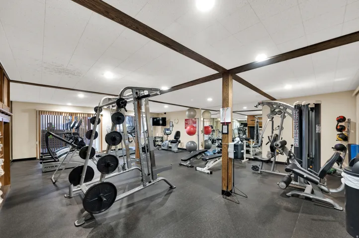 Dedicated fitness area featuring rubber flooring, exposed wood beams, track lighting, and substantial exercise equipment