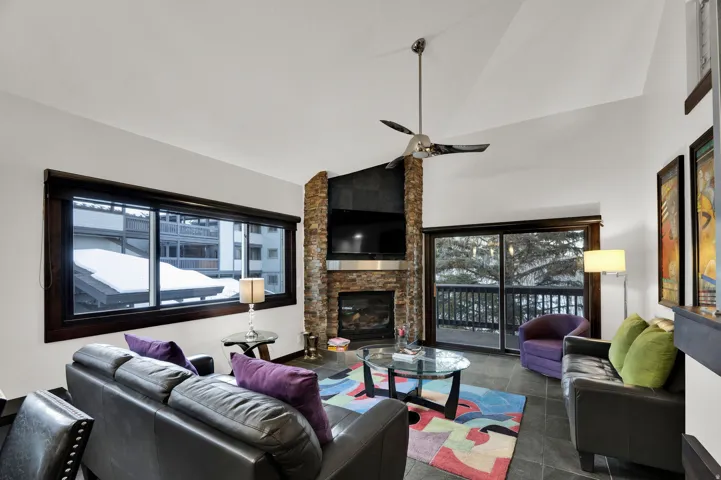 Vaulted ceiling with ceiling fan, stone-clad fireplace, large sliding glass door, dark-framed window, and stone-finish flooring