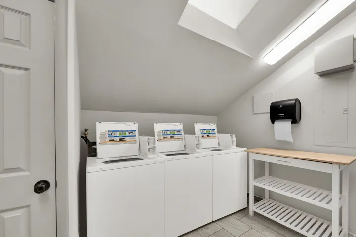 Laundry facility with three top-loading washing machines, a wood-finish utility table, and overhead fluorescent lighting