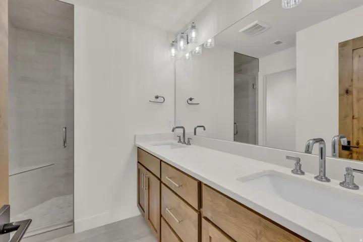 Dual sink vanity with wood-finish cabinetry, white countertop, chrome fixtures, and a frameless glass shower enclosure