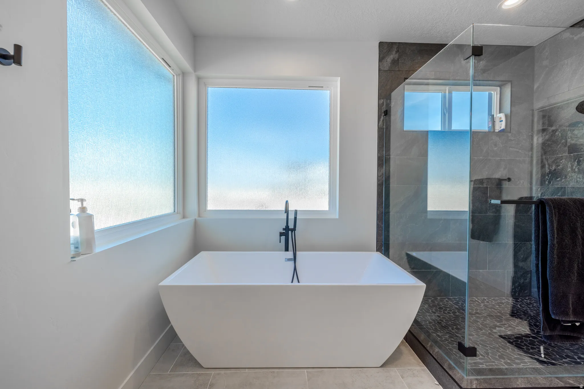 Freestanding soaking tub with matte black fixtures, glass-enclosed shower with dark tile surround, and light tile flooring