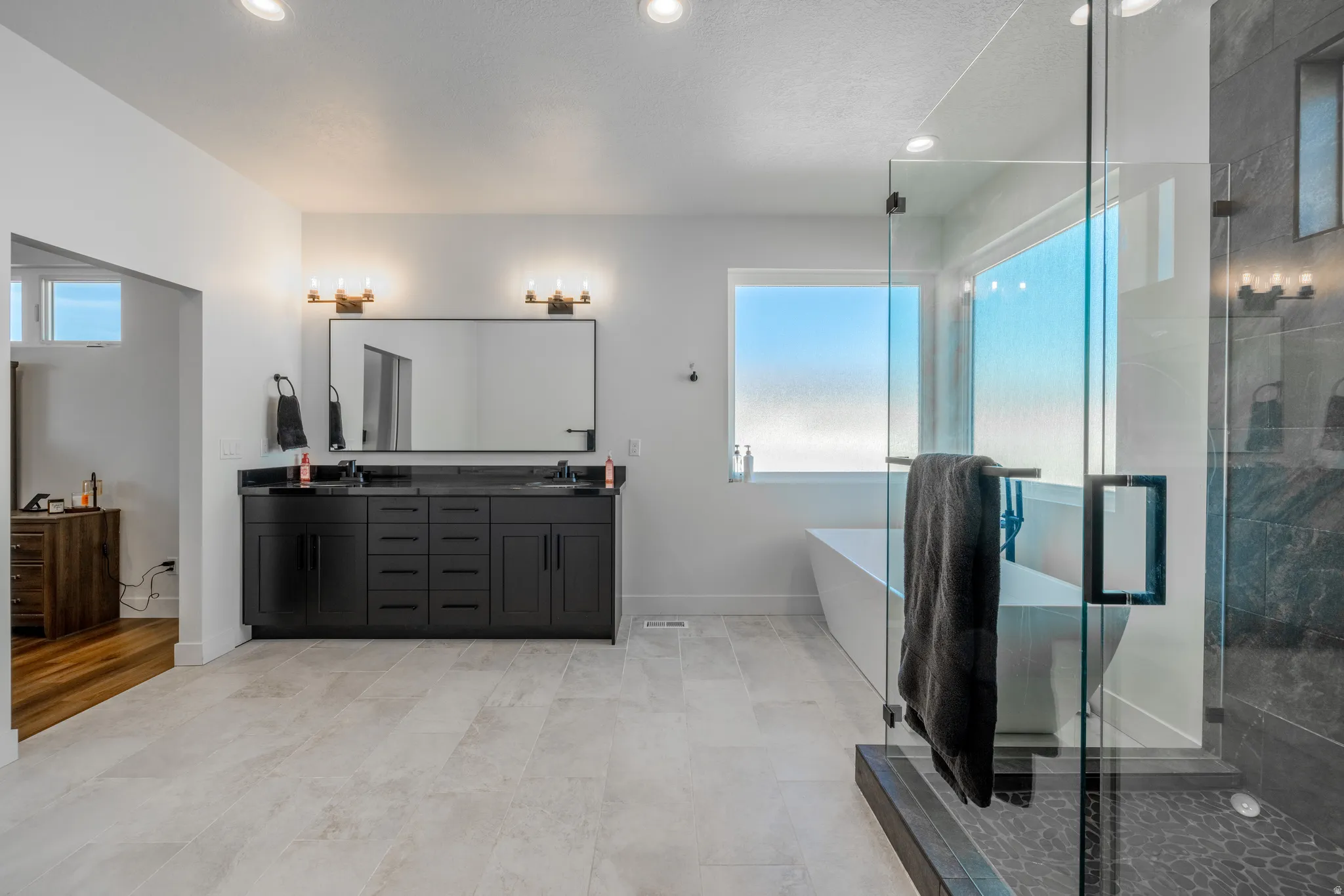 Dual vanity with dark cabinetry and black countertop, freestanding soaking tub, glass-enclosed shower, light-toned tile flooring, and recessed lighting