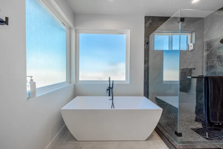 Freestanding soaking tub with matte black fixtures, glass-enclosed shower with dark tile surround, and light tile flooring