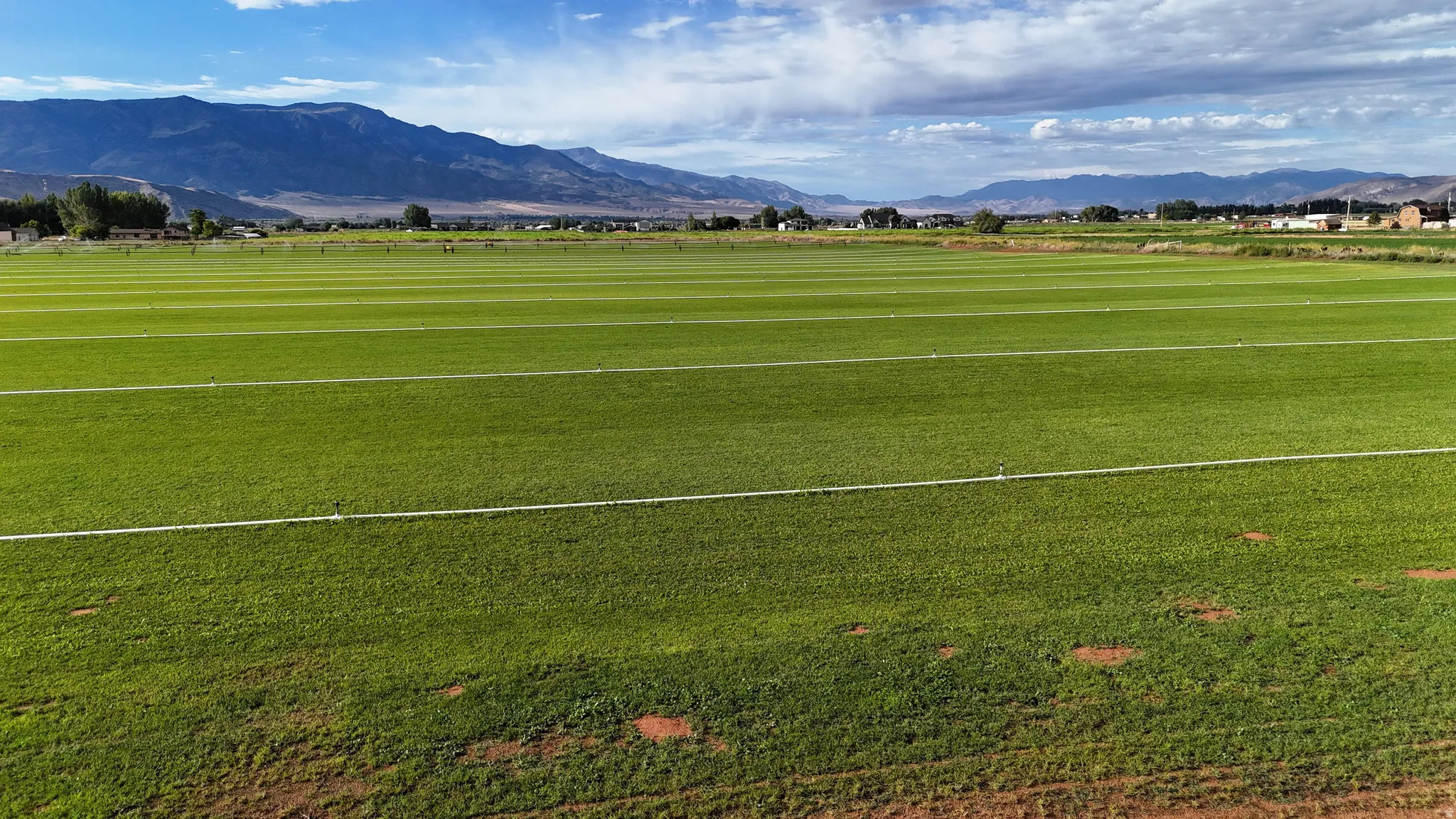 Expansive green field with visible irrigation lines, set against a backdrop of distant mountains under a partly cloudy sky