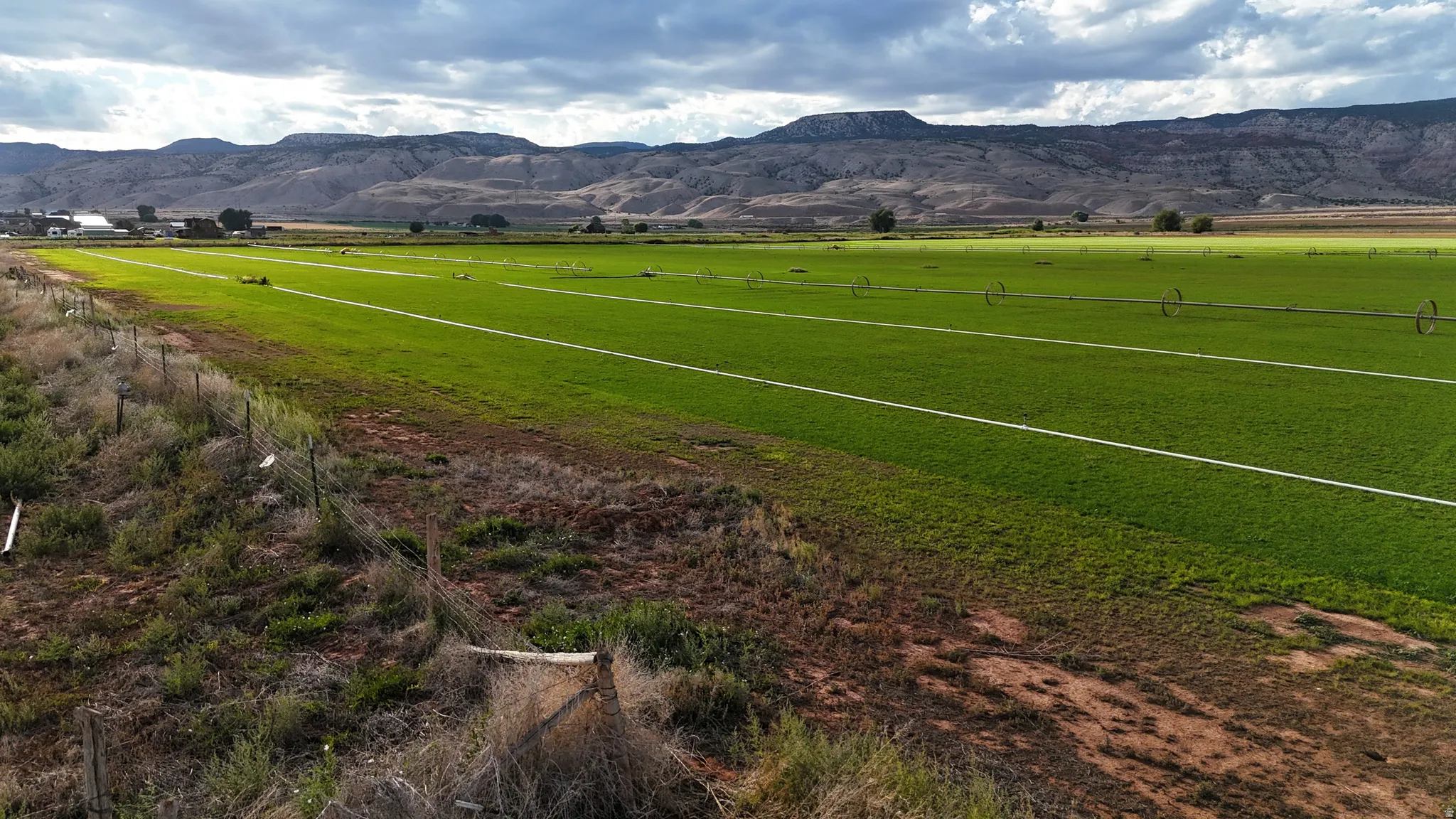 Expansive agricultural land featuring a verdant crop and multiple irrigation systems, bordered by a wire fence, with distant mountains providing a scenic backdrop