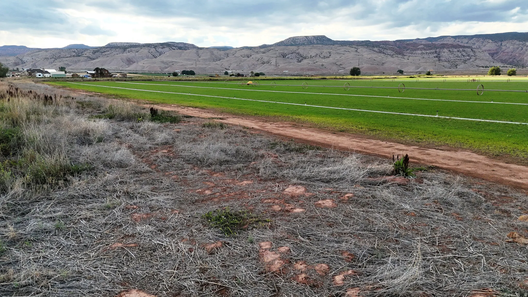 Expansive undeveloped land with a dirt path, bordered by green agricultural fields and distant mountainous terrain