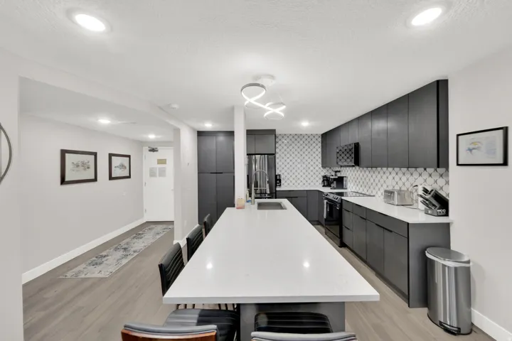 Open-concept kitchen featuring an elongated island with a light-toned countertop, dark-toned cabinetry, a geometric pattern backsplash, and wood-finish flooring