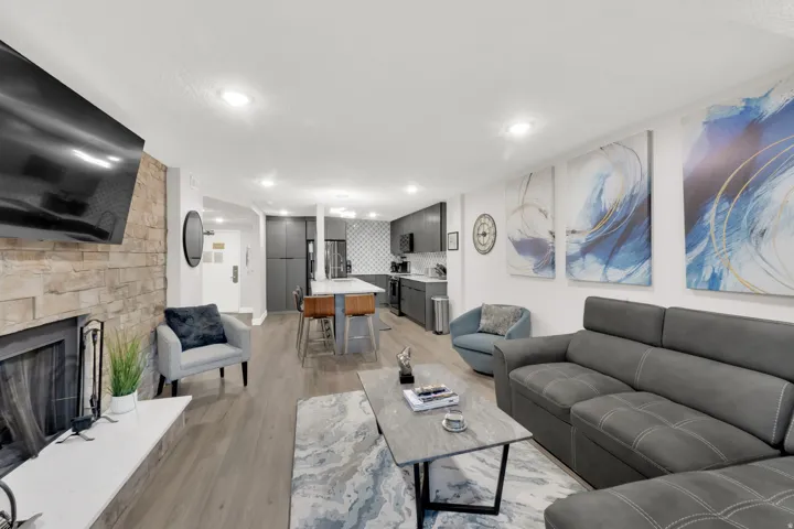 Fireplace with stone surround and white mantel, wood-finish flooring, recessed lighting, and a kitchen featuring dark flat-panel cabinetry and stainless steel appliances