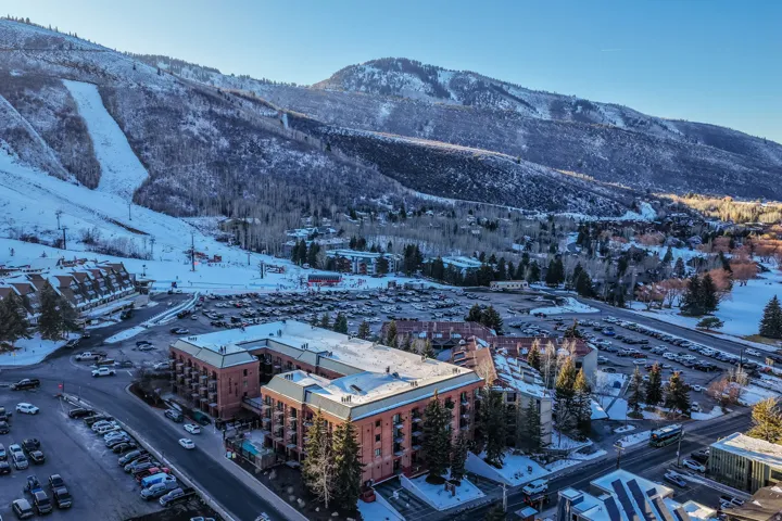 Mountain backdrop with snow-covered terrain, surrounding mature trees, multi-level buildings with balconies, and multiple parking areas