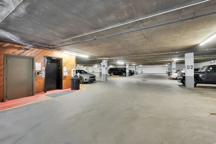 Covered parking garage with concrete flooring and ceiling, illuminated by overhead lighting, featuring marked parking spaces and clear structural columns
