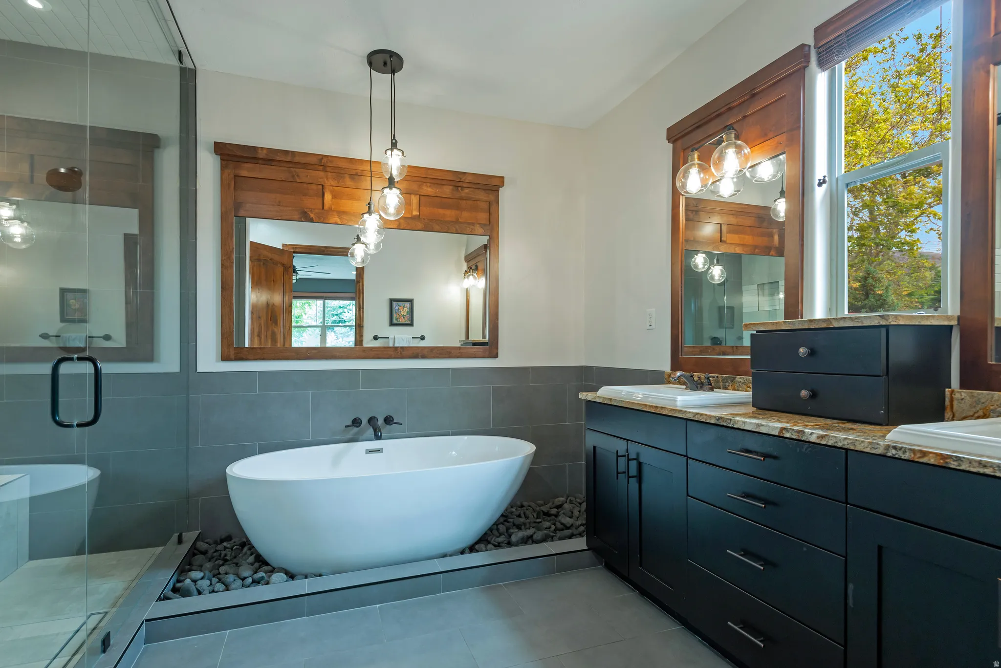 Freestanding soaking tub with river rock surround, dual vanity with granite countertops, wood-framed mirrors, and a glass-enclosed shower