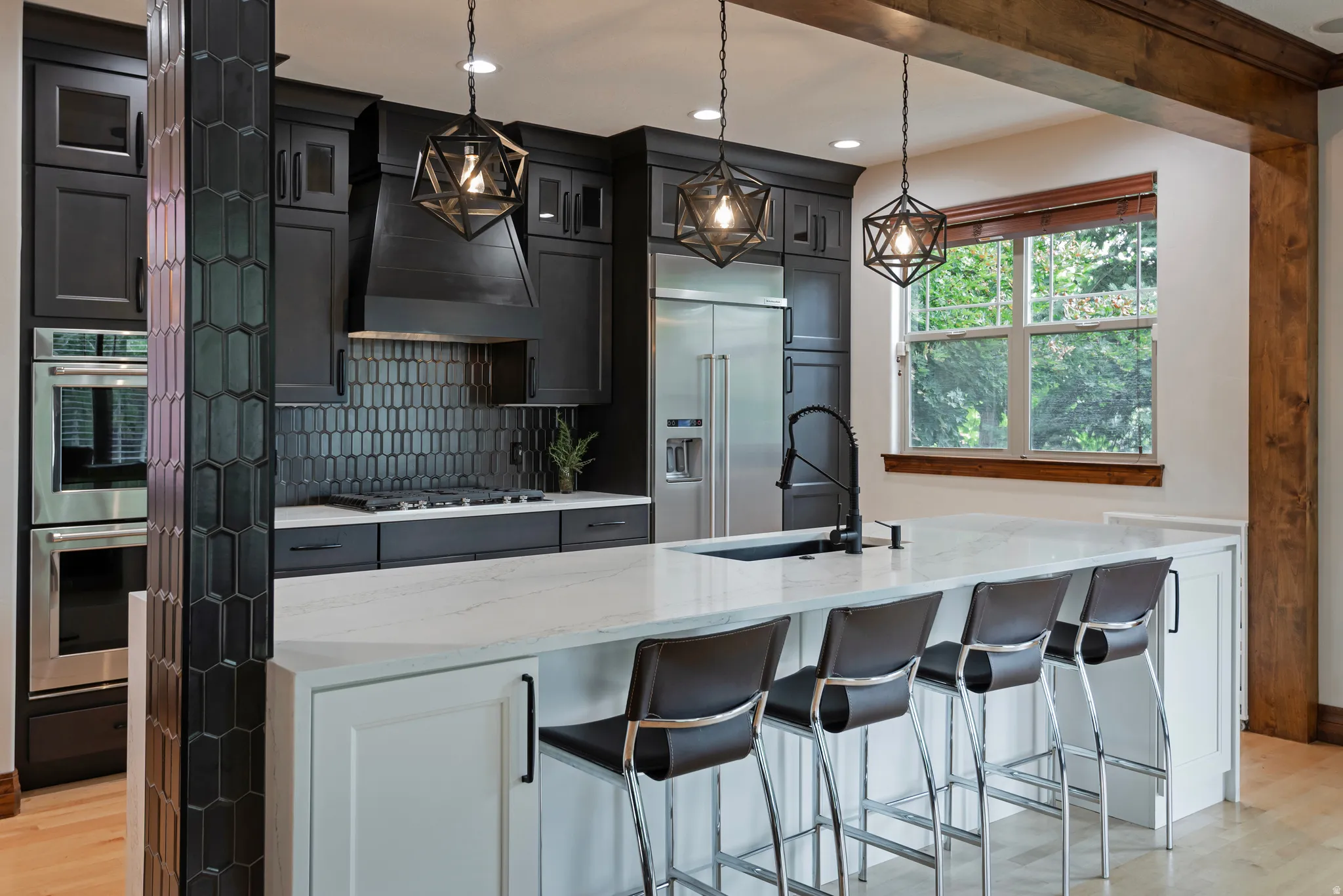 Sleek kitchen featuring dark cabinetry, stainless steel appliances, a white countertop island with seating, a black tile backsplash, and decorative pendant lighting