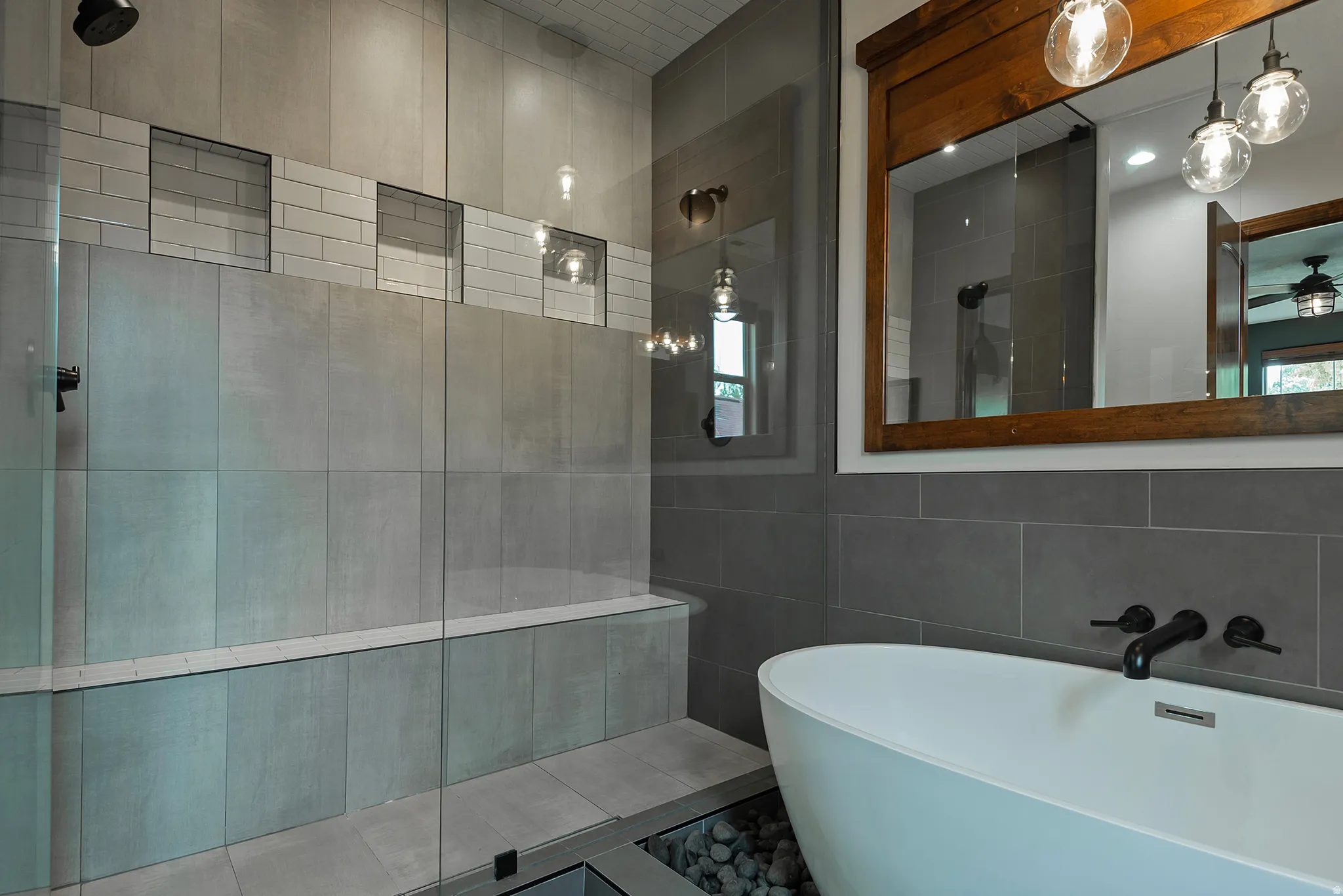 Freestanding soaking tub with floor-level river rock detailing, large format tile walls, subway tile accents, and a wide wooden-framed mirror with pendant lighting