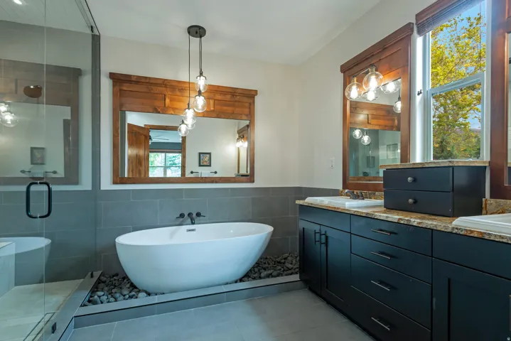 Freestanding soaking tub with river rock surround, dual vanity with granite countertops, wood-framed mirrors, and a glass-enclosed shower
