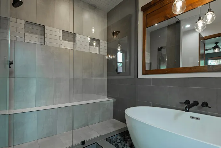 Freestanding soaking tub with floor-level river rock detailing, large format tile walls, subway tile accents, and a wide wooden-framed mirror with pendant lighting