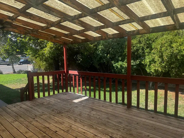 Wood deck with a slatted railing, covered by a wood-framed pergola with a translucent roof, overlooking a grassy area with mature foliage