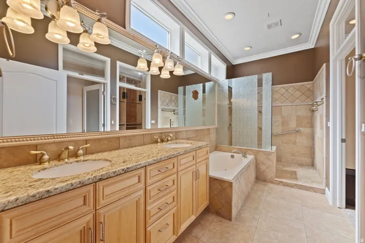 Dual vanity with stone countertop, tile flooring, walk-in shower with textured glass panels, jetted soaking tub, and transom windows