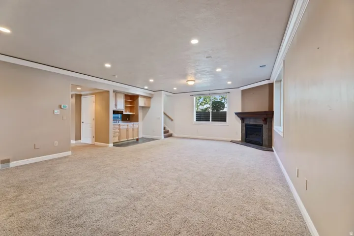 Spacious carpeted room featuring recessed lighting, a built-in wet bar with cabinetry, a corner fireplace with a dark wood mantel, and multiple windows with horizontal blinds
