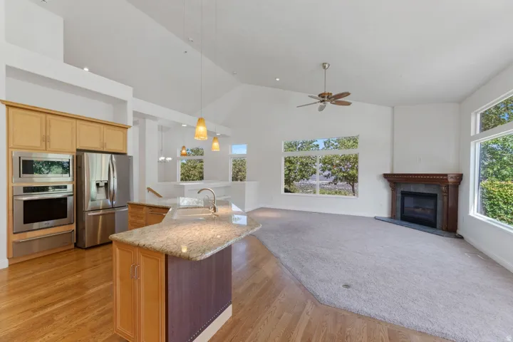 Open-concept interior featuring vaulted ceilings, wood-finish flooring, a kitchen island with stone countertops, stainless steel appliances, and a fireplace with a decorative mantel