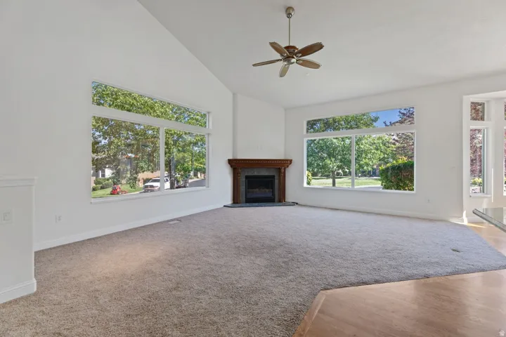 Vaulted ceiling with a ceiling fan, fireplace with a wood mantel, large windows, and carpet flooring transitioning to wood-finish flooring