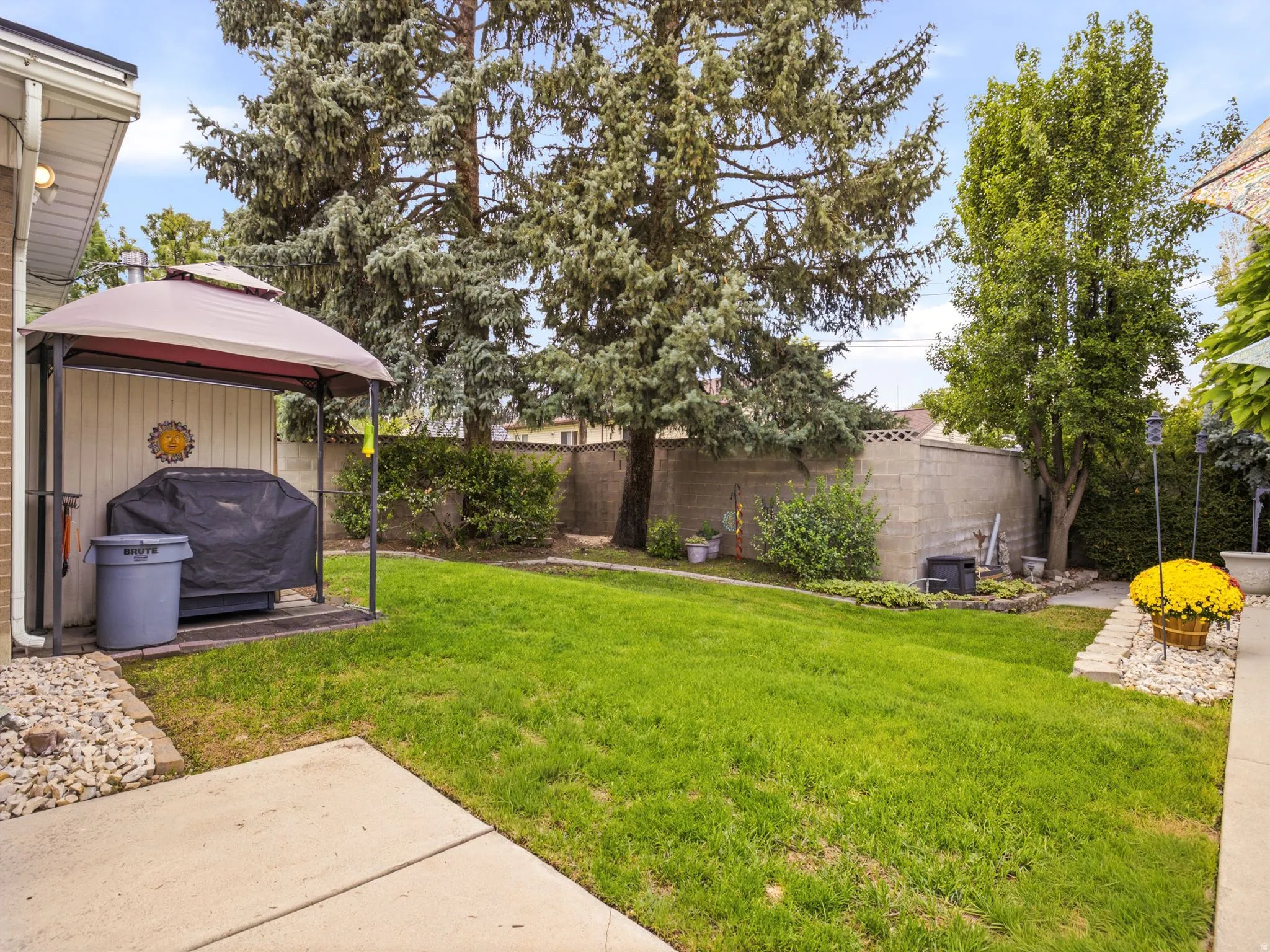 Spacious backyard oasis featuring a manicured lawn, mature evergreen and deciduous trees, concrete patio, detached storage shed with peaked roof and protective cover, decorative gravel landscaping, and perimeter fencing