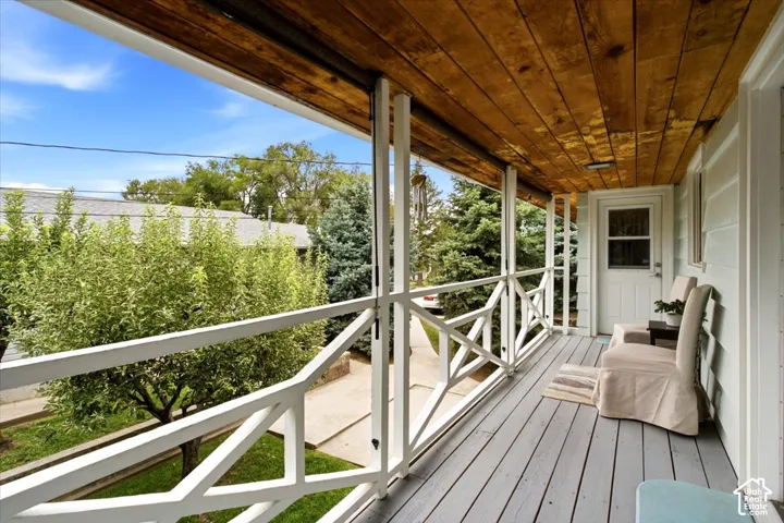 Covered porch with wood plank ceiling, light gray wood-finish flooring, white railing with X-pattern detailing, and white paneled door with window