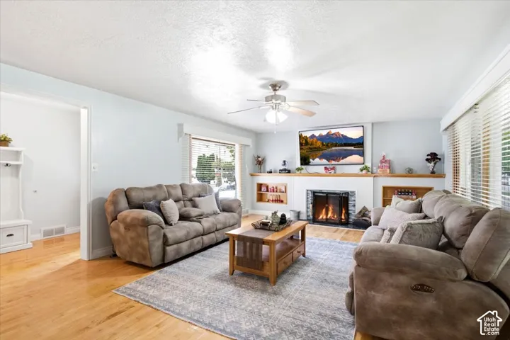 Hardwood flooring, fireplace with stone surround, built-in shelving, white window blinds, and a ceiling fan
