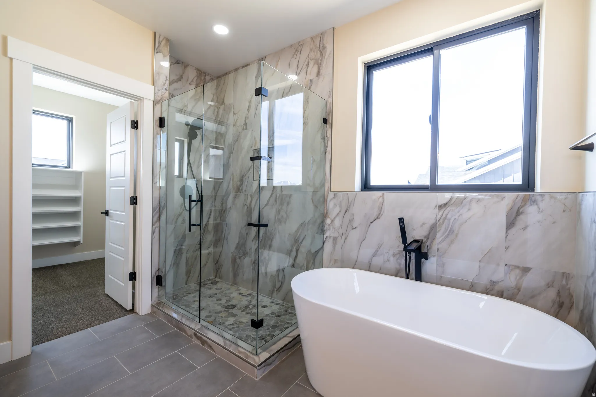 Freestanding bathtub with matte black floor-mounted faucet, glass-enclosed shower with stone-finish tile surround, and neutral tile flooring