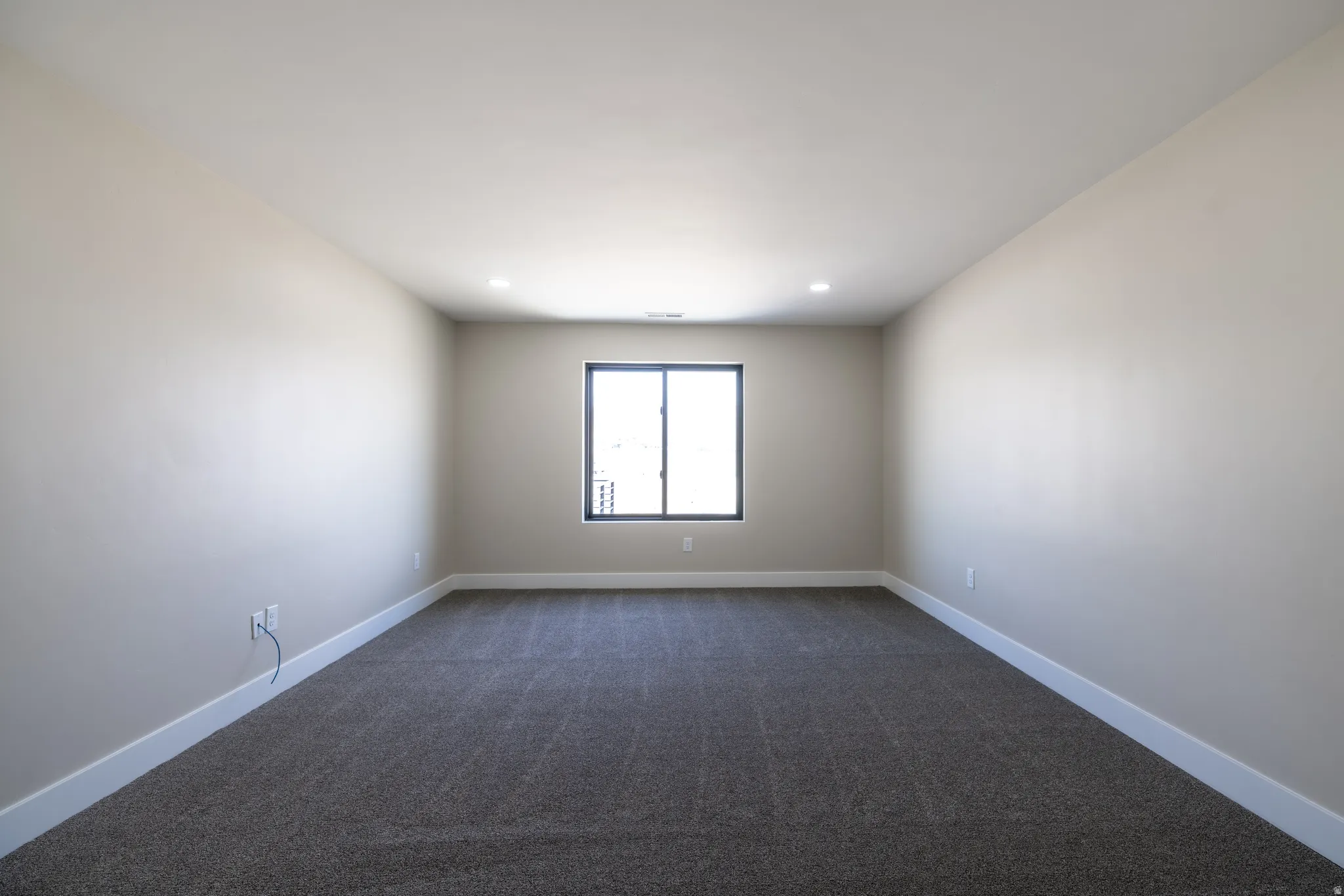 Expansive room with neutral tone walls, dark carpet, recessed lighting, and a single window with dark framing